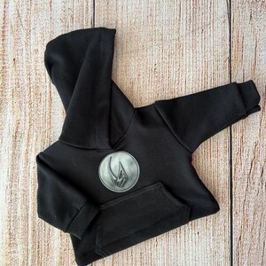 Op de afbeelding: Een effen zwarte hoodie met lange mouwen en een ronde grijze patch op de borst. De patch toont een verhoogde, metaalkleurige afbeelding van een gestileerde klauw- of hoornvorm. De hoodie is voorzien van een kangoeroezak en geribbelde boorden.