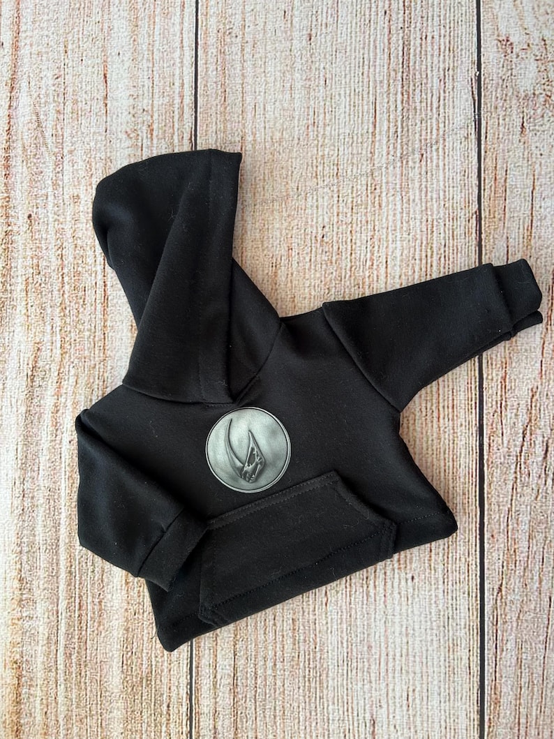 Op de afbeelding: Een effen zwarte hoodie met lange mouwen en een ronde grijze patch op de borst. De patch toont een verhoogde, metaalkleurige afbeelding van een gestileerde klauw- of hoornvorm. De hoodie is voorzien van een kangoeroezak en geribbelde boorden.