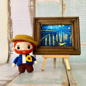 Puede incluir: Una muñeca de ganchillo con pelo rojo, barba y sombrero amarillo, que lleva una chaqueta azul con un broche de girasol, junto a un pequeño cuadro enmarcado de un paisaje nocturno con reflejos de luz amarilla sobre el agua, en un caballete.