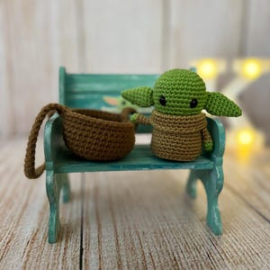 May include: A small green crocheted character with large eyes and brown crocheted clothing sitting on a teal bench with a brown crocheted basket beside it.