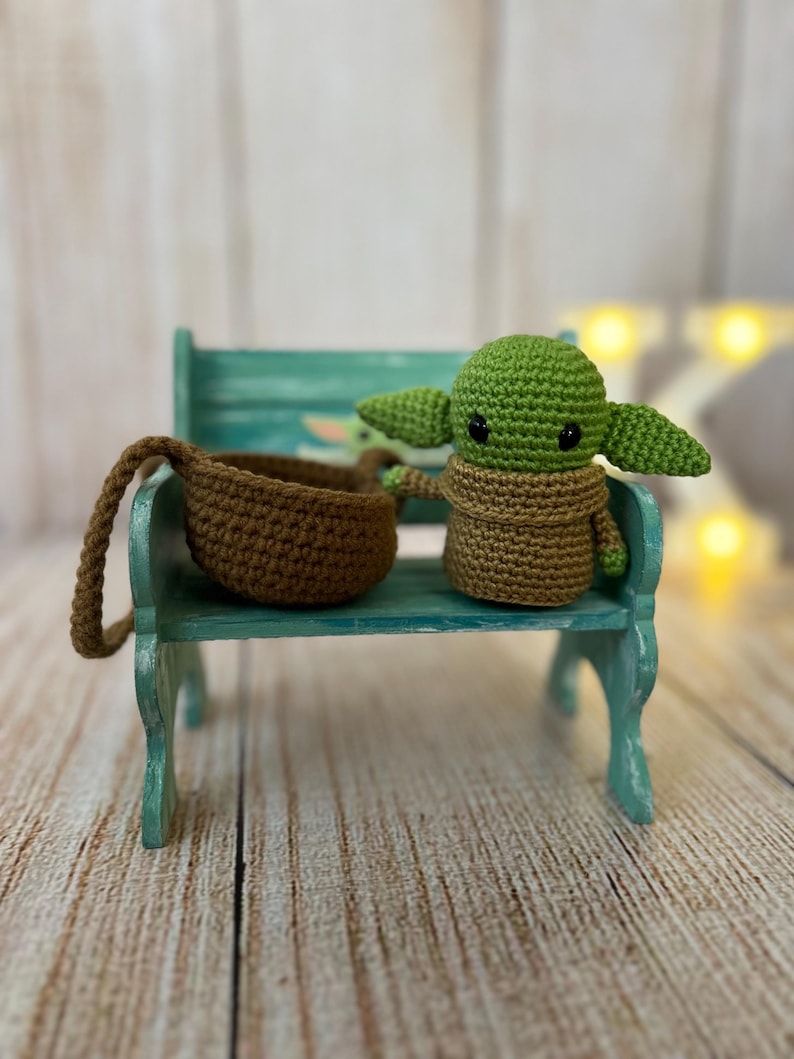 May include: A small green crocheted character with large eyes and brown crocheted clothing sitting on a teal bench with a brown crocheted basket beside it.