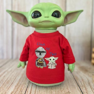 Puede incluir: Una figura de criatura verde con grandes ojos negros y orejas puntiagudas que lleva una camiseta roja de manga larga. La camiseta presenta un gráfico bordado de un personaje con casco y una pequeña criatura verde con el texto azul Clan of Two.