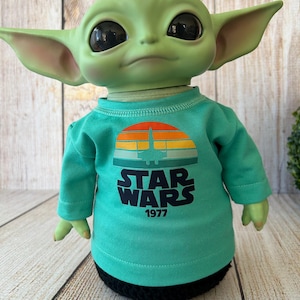 Peut inclure: Une poup&eacute;e verte avec de grands yeux noirs et des oreilles pointues, portant un t-shirt turquoise &agrave; manches longues. Le t-shirt pr&eacute;sente un motif de coucher de soleil r&eacute;tro avec un vaisseau spatial et les mots "STAR WARS 1977" en blanc.