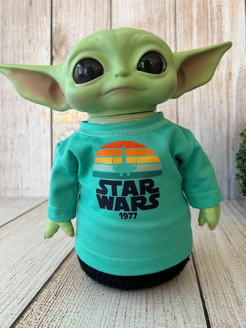 Peut inclure: Une poup&eacute;e verte avec de grands yeux noirs et des oreilles pointues, portant un t-shirt turquoise &agrave; manches longues. Le t-shirt pr&eacute;sente un motif de coucher de soleil r&eacute;tro avec un vaisseau spatial et les mots "STAR WARS 1977" en blanc.