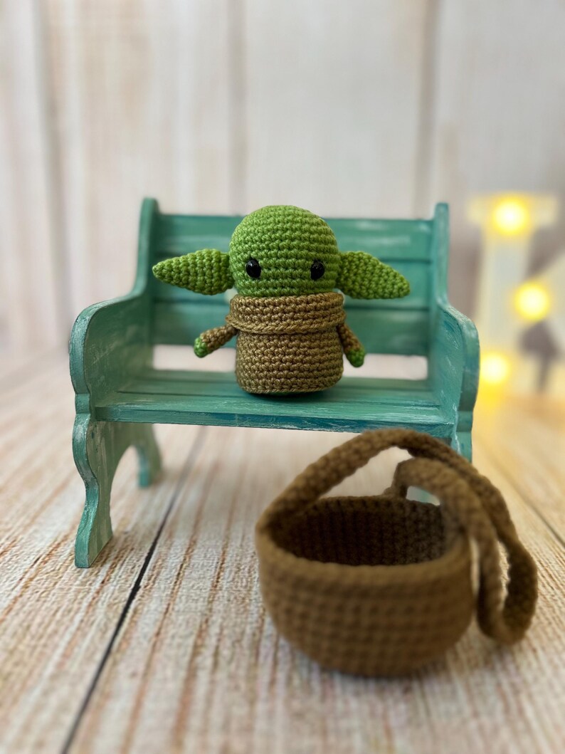 May include: A small green crocheted character with large ears and brown eyes sits on a teal bench. A brown crocheted basket sits on the wooden floor.