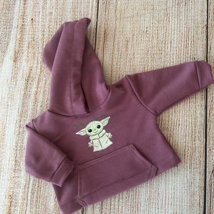 Op de afbeelding: Een mauvekleurige hoodie met lange mouwen en een afbeelding van een groen personage met grote oren, met de tekst "This is the way" erboven gedrukt. Het kledingstuk is voorzien van een kangoeroezak aan de voorzijde, geribbelde boorden en een capuchon.