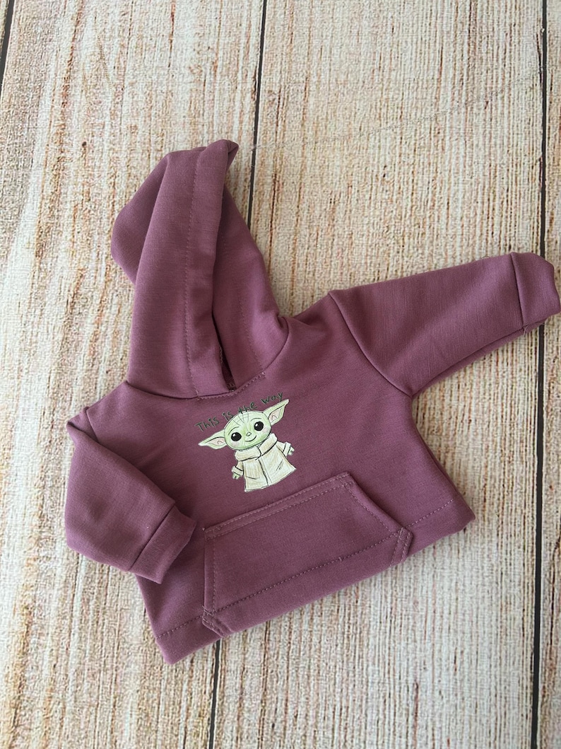 Op de afbeelding: Een mauvekleurige hoodie met lange mouwen en een afbeelding van een groen personage met grote oren, met de tekst "This is the way" erboven gedrukt. Het kledingstuk is voorzien van een kangoeroezak aan de voorzijde, geribbelde boorden en een capuchon.