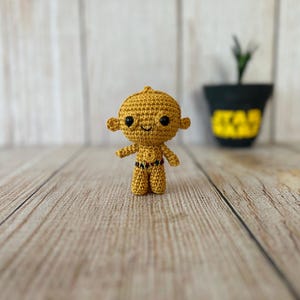 以下が含まれることがあります： 黄色い体、黒い目、小さな黒いベルトが付いた、かぎ針編みのおもちゃのロボットのフィギュア。おもちゃは木製の表面に立っています。