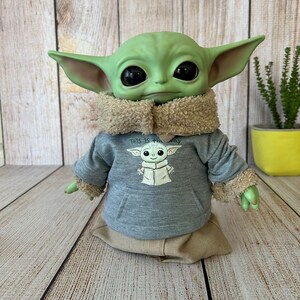 May include: A green Grogu doll wearing a gray hoodie with a Grogu graphic and the text "THIS IS THE WAY". The doll has large black eyes, pointed ears, and a tan fur-lined collar and cuffs. It is sitting on a tan fabric base.