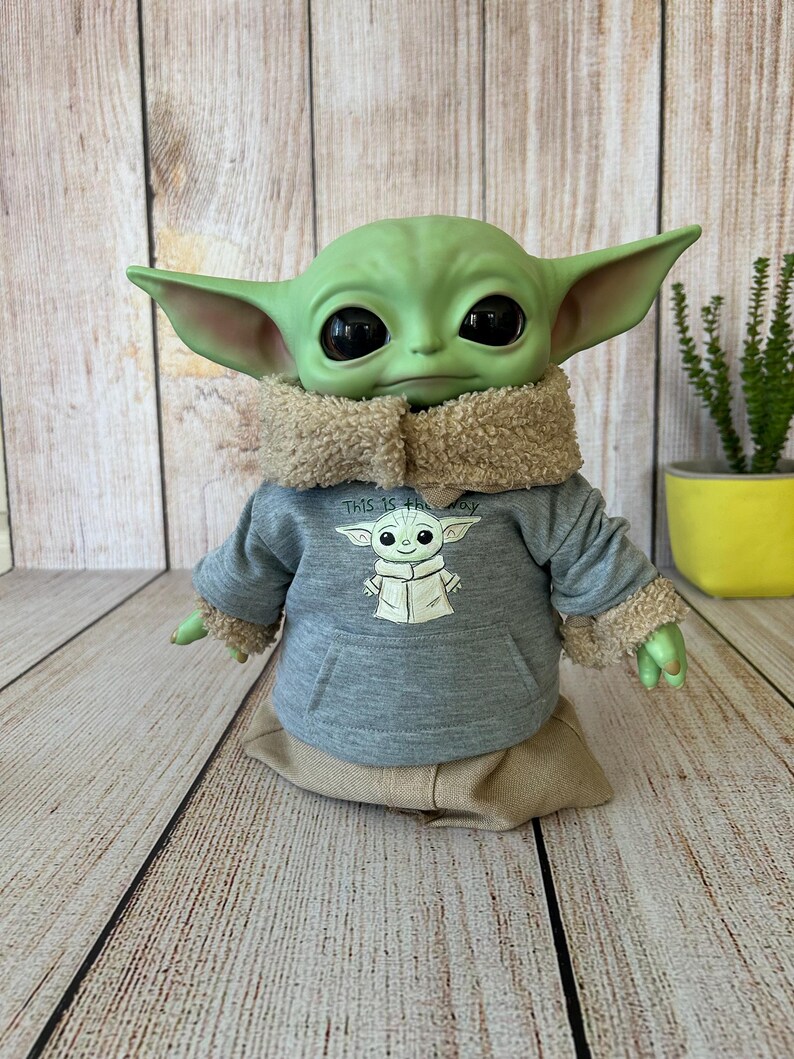 May include: A green Grogu doll wearing a gray hoodie with a Grogu graphic and the text "THIS IS THE WAY". The doll has large black eyes, pointed ears, and a tan fur-lined collar and cuffs. It is sitting on a tan fabric base.