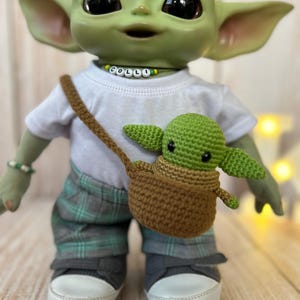 May include: A green plush toy with large ears and big eyes wearing a white shirt with the word "COLLI" spelled out in colorful beads, gray and green plaid pants, and gray sneakers. The toy is holding a smaller green plush toy in a brown crocheted basket.
