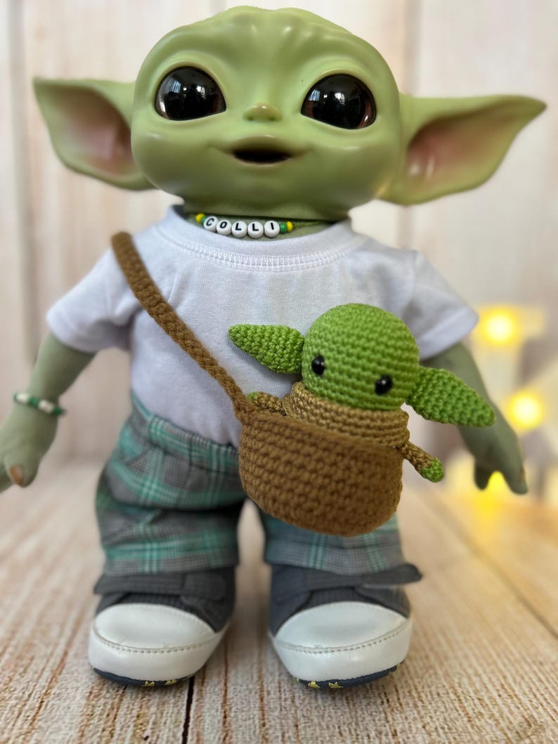 May include: A green plush toy with large ears and big eyes wearing a white shirt with the word "COLLI" spelled out in colorful beads, gray and green plaid pants, and gray sneakers. The toy is holding a smaller green plush toy in a brown crocheted basket.