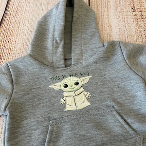 May include: A gray hooded sweatshirt featuring a front pocket and a hood. The sweatshirt has an illustration of a green alien character with the text "This is the Way" above the character.