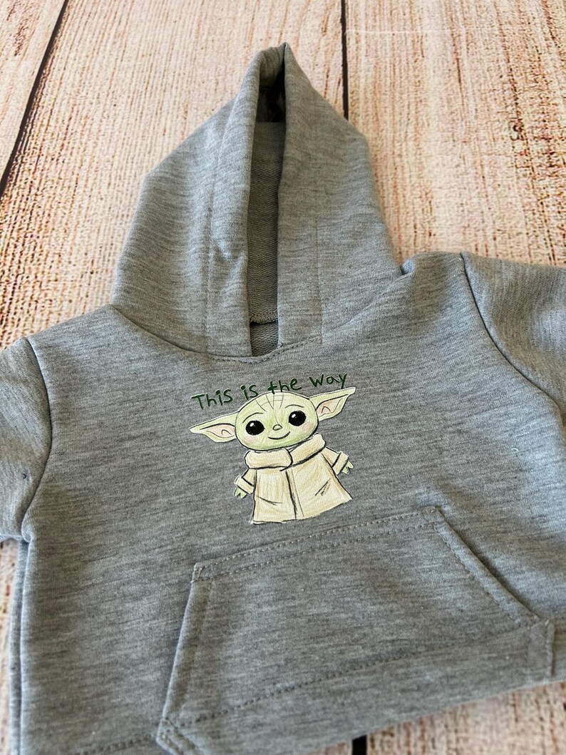 May include: A gray hooded sweatshirt featuring a front pocket and a hood. The sweatshirt has an illustration of a green alien character with the text "This is the Way" above the character.