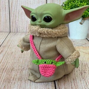 K&ouml;nnte beinhalten: Eine gr&uuml;n-rosa geh&auml;kelte Handtasche mit einem gr&uuml;nen Charaktergesicht auf der Vorderseite. Die Tasche wird als Umh&auml;ngetasche von einem gr&uuml;nen Pl&uuml;schtier getragen.