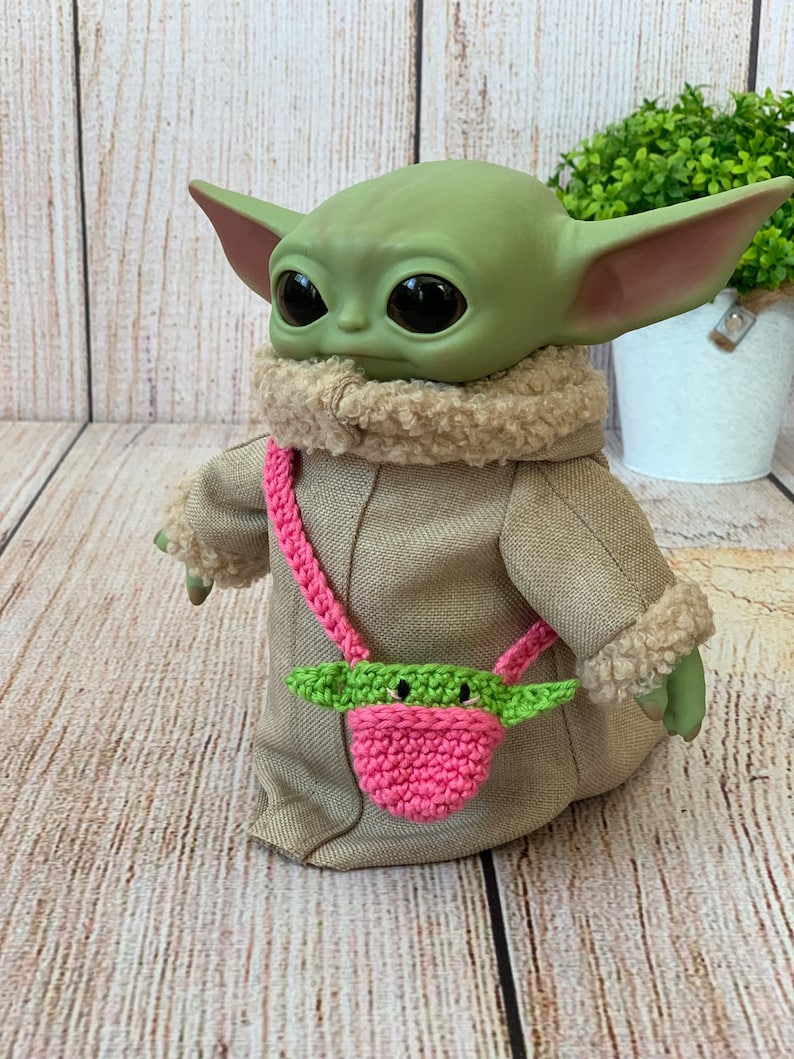 K&ouml;nnte beinhalten: Eine gr&uuml;n-rosa geh&auml;kelte Handtasche mit einem gr&uuml;nen Charaktergesicht auf der Vorderseite. Die Tasche wird als Umh&auml;ngetasche von einem gr&uuml;nen Pl&uuml;schtier getragen.