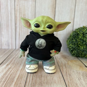 Op de afbeelding: Een groenhuidige wezenpop met grote donkere ogen en puntige oren. Het draagt een zwarte hoodie met een cirkelvormig zilveren embleem op de borst, een groen-grijze geruite broek en witte sneakers met een grijze rand langs de zolen.