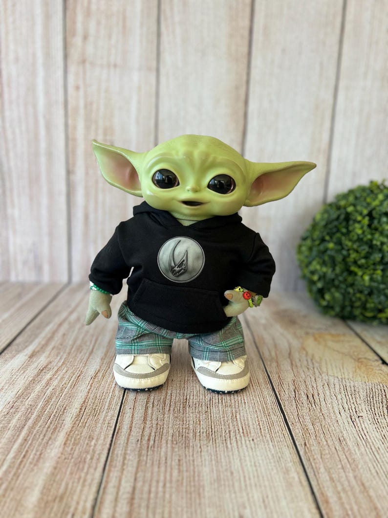 Op de afbeelding: Een groenhuidige wezenpop met grote donkere ogen en puntige oren. Het draagt een zwarte hoodie met een cirkelvormig zilveren embleem op de borst, een groen-grijze geruite broek en witte sneakers met een grijze rand langs de zolen.