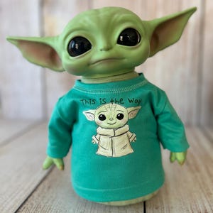Peut inclure: Poup&eacute;e verte repr&eacute;sentant un personnage avec de grands yeux noirs et des oreilles pointues, portant un t-shirt vert d'eau &agrave; manches longues. Le t-shirt pr&eacute;sente une image de dessin anim&eacute; du personnage et le texte "This is the Way".