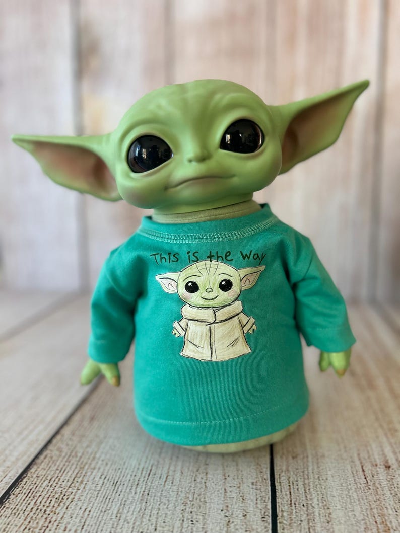 Peut inclure: Poup&eacute;e verte repr&eacute;sentant un personnage avec de grands yeux noirs et des oreilles pointues, portant un t-shirt vert d'eau &agrave; manches longues. Le t-shirt pr&eacute;sente une image de dessin anim&eacute; du personnage et le texte "This is the Way".