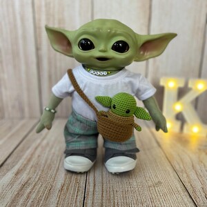 May include: A green plush toy dressed in a white t-shirt with the word "COULD" on it, plaid pants, and sneakers. The toy is holding a small green plush toy in a brown crocheted bag.