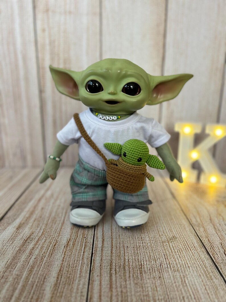 May include: A green plush toy dressed in a white t-shirt with the word "COULD" on it, plaid pants, and sneakers. The toy is holding a small green plush toy in a brown crocheted bag.