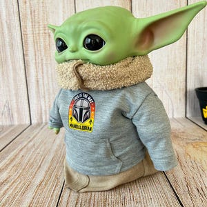 May include: A green Grogu figure wearing a gray hoodie with a "This is the Way" Mandalorian logo. The figure has large black eyes, large ears, and a tan scarf. The figure is wearing tan pants.
