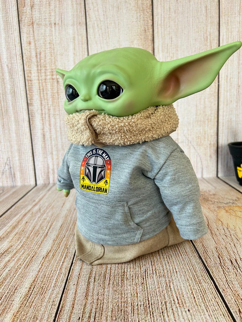 May include: A green Grogu figure wearing a gray hoodie with a "This is the Way" Mandalorian logo. The figure has large black eyes, large ears, and a tan scarf. The figure is wearing tan pants.