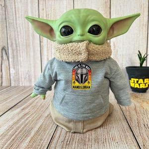 May include: A green Grogu doll wearing a gray hoodie with the text "This is the Way" and a Mandalorian logo. The doll has large black eyes and tan clothing. A small Star Wars pot with a plant is in the background.