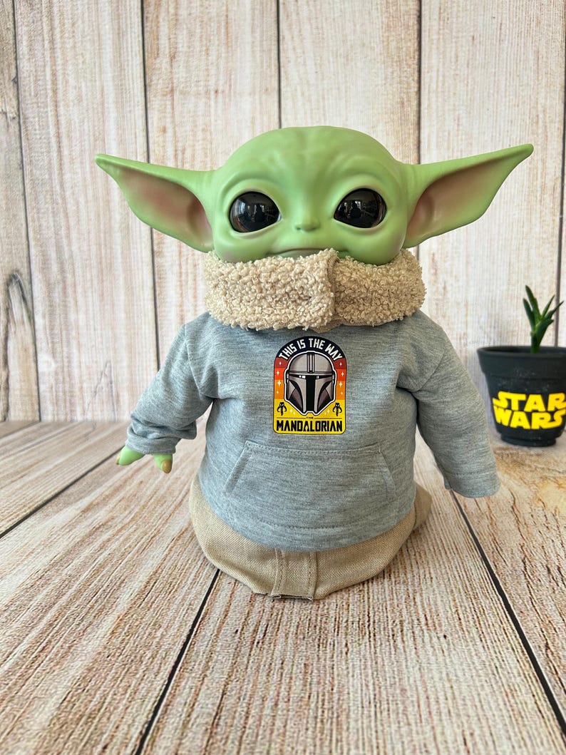 May include: A green Grogu doll wearing a gray hoodie with the text "This is the Way" and a Mandalorian logo. The doll has large black eyes and tan clothing. A small Star Wars pot with a plant is in the background.