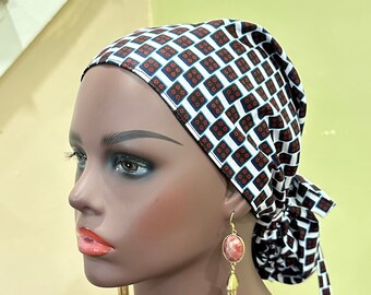 May include: A patterned headscarf with a repeating geometric design in white, navy, and red. The head covering is tied at the back. The mannequin is wearing gold earrings with a pink stone and gold tassels.
