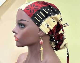 May include: A beige, red, and black patterned headscarf with a geometric design. The headscarf is tied in a knot at the back of the head. The person wearing the headscarf is also wearing long, gold earrings with a pink stone.