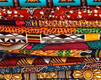 May include: A stack of colorful African wax print fabrics. The fabrics feature various patterns, including geometric shapes, floral designs, and abstract motifs. Colors include blue, red, orange, yellow, and black. The fabrics are neatly folded, showcasing the diverse designs.