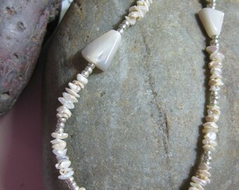 May include: A white shell necklace with silver beads. The necklace is made of irregular shaped white shell pieces and silver beads. The necklace is lying on a gray rock.