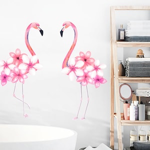 May include: Two pink flamingo wall decals with pink flowers. The decals are on a white wall in a bathroom with a white bathtub and a wooden shelf unit with towels and toiletries.