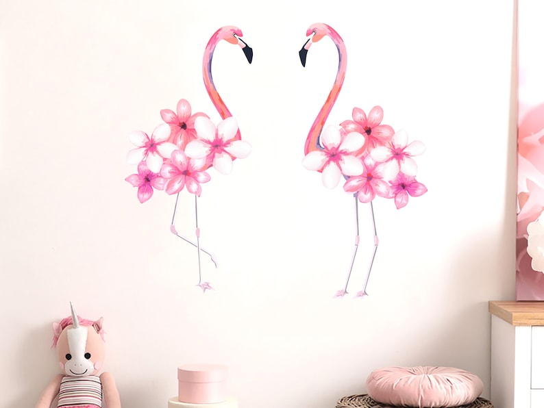 May include: Two pink flamingos with pink flowers on their backs. The flamingos are standing on a white background.