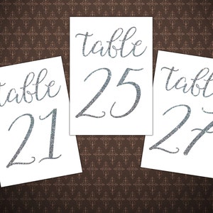 May include: Three white table number cards with silver glitter lettering. The cards read "table 21", "table 25", and "table 27".