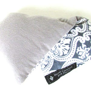 Puede incluir: Dos paquetes de arroz. Uno es de forma rectangular gris claro. El otro es de forma triangular con un estampado floral en blanco y negro. Una etiqueta negra dice "Divine Comfort Rice Packs."
