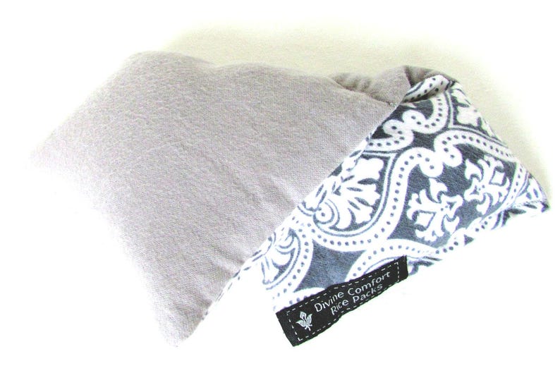 Puede incluir: Dos paquetes de arroz. Uno es de forma rectangular gris claro. El otro es de forma triangular con un estampado floral en blanco y negro. Una etiqueta negra dice "Divine Comfort Rice Packs."