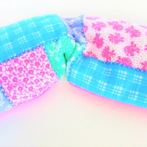 May include: A patchwork rice pack in shades of blue, pink, and green. The fabric features floral and plaid patterns, stitched together with blue thread. A black label reads "Divine Comfort Rice Packs."