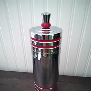 May include: A tall, cylindrical, chrome-finished container with red accent bands. The container has a matching lid and a small, conical top. The design is sleek and modern, suitable for kitchen or bar decor.