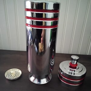 May include: A shiny, chrome cocktail shaker with red bands and a matching lid. The shaker is cylindrical with a flared base, and a strainer and lid are next to it. The reflective surface shows a distorted reflection.
