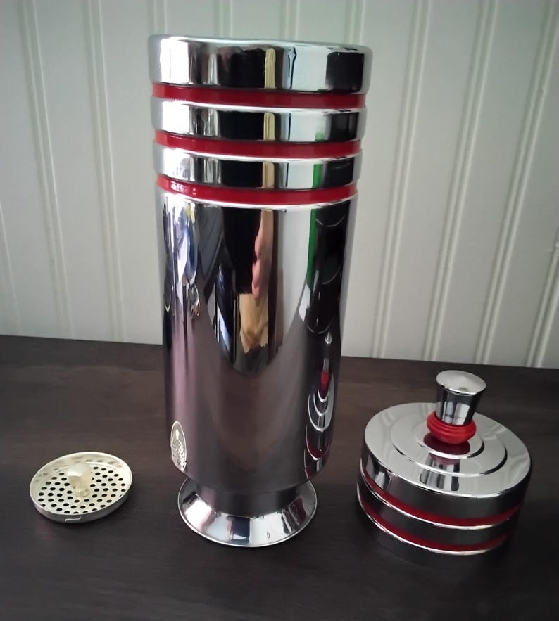 May include: A shiny, chrome cocktail shaker with red bands and a matching lid. The shaker is cylindrical with a flared base, and a strainer and lid are next to it. The reflective surface shows a distorted reflection.