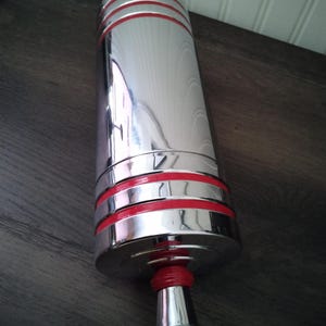 May include: A cylindrical, chrome-finished object with three red bands near the top and one at the base. The object has a flared, silver base with a red rubber ring. The reflective surface suggests a barware item.