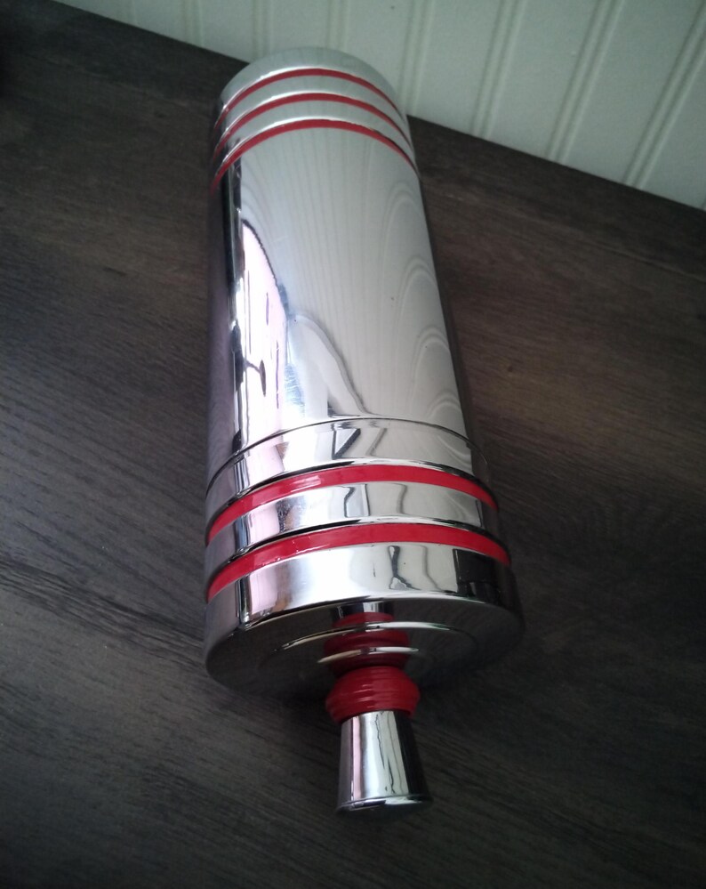 May include: A cylindrical, chrome-finished object with three red bands near the top and one at the base. The object has a flared, silver base with a red rubber ring. The reflective surface suggests a barware item.