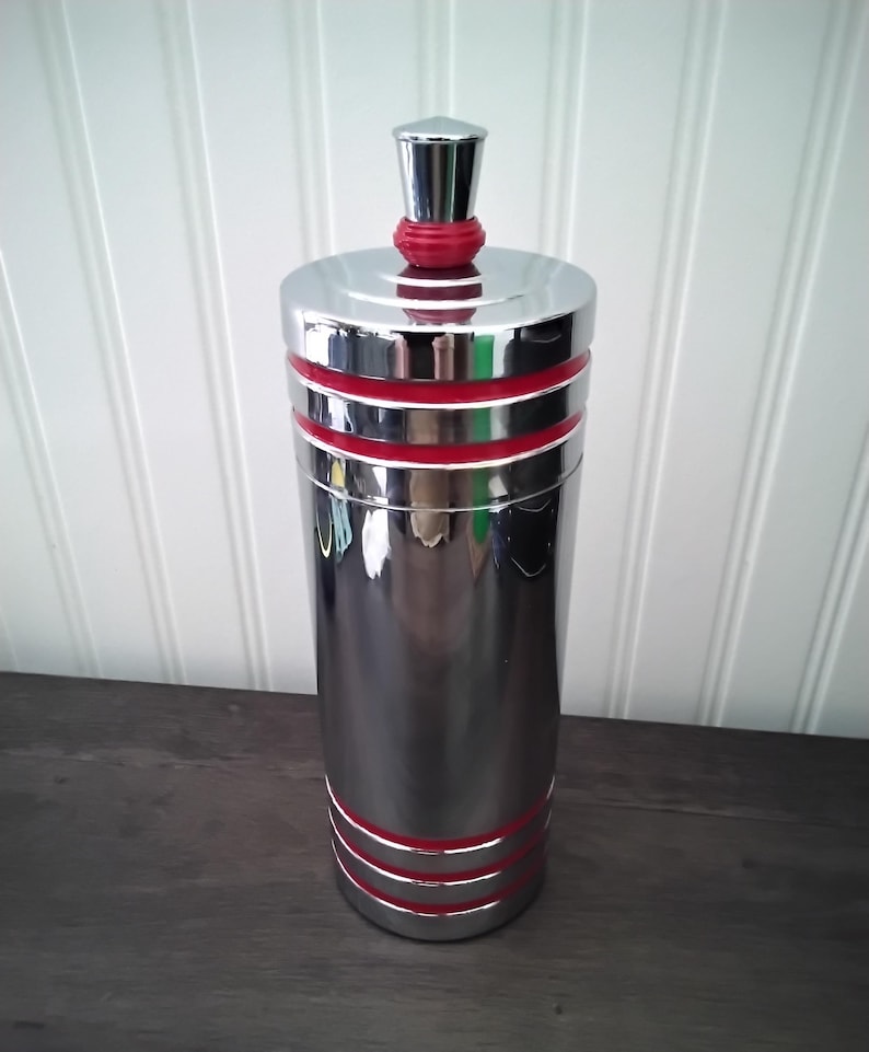 May include: A tall, cylindrical, chrome-finished container with red accent bands. The container has a matching lid and a small, conical top. The design is sleek and modern, suitable for kitchen or bar decor.
