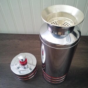 May include: A vintage chrome cocktail shaker with a lid and strainer. The shaker has red accent stripes and a red top on the lid. The shaker is on a dark wooden surface.