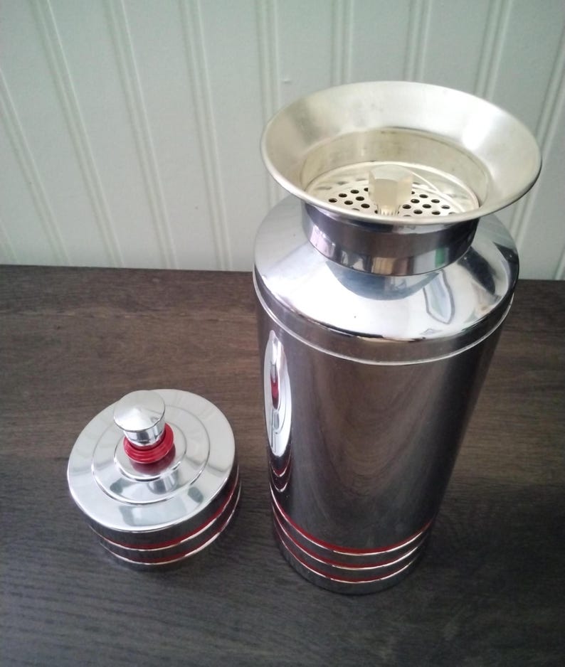 May include: A vintage chrome cocktail shaker with a lid and strainer. The shaker has red accent stripes and a red top on the lid. The shaker is on a dark wooden surface.