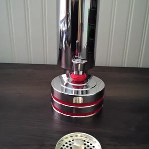 May include: A chrome-plated kitchen sink strainer and a matching cylindrical object with red accents. The cylindrical object has a base and a top, with a red band around the middle. The strainer is round with small holes.