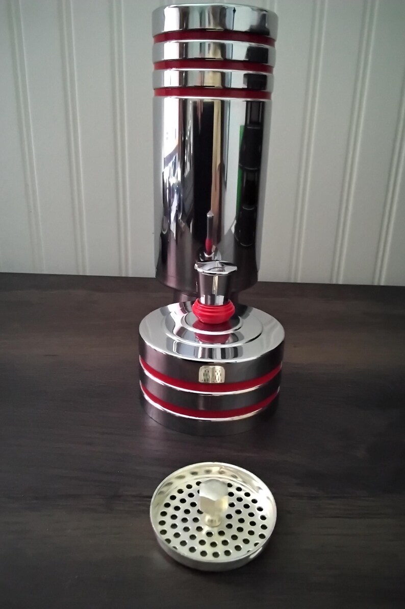 May include: A chrome-plated kitchen sink strainer and a matching cylindrical object with red accents. The cylindrical object has a base and a top, with a red band around the middle. The strainer is round with small holes.